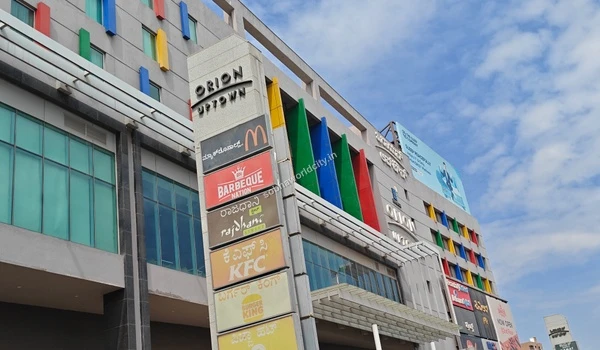 Wide street-level perspective of the shopping center's modern architecture with colorful window panels and a large Orion Uptown logo near Sobha World City