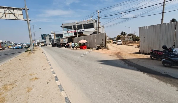 A wide perspective of the traffic and commercial vehicles on Old Madras Road approaching the Sobha World City site entrance.